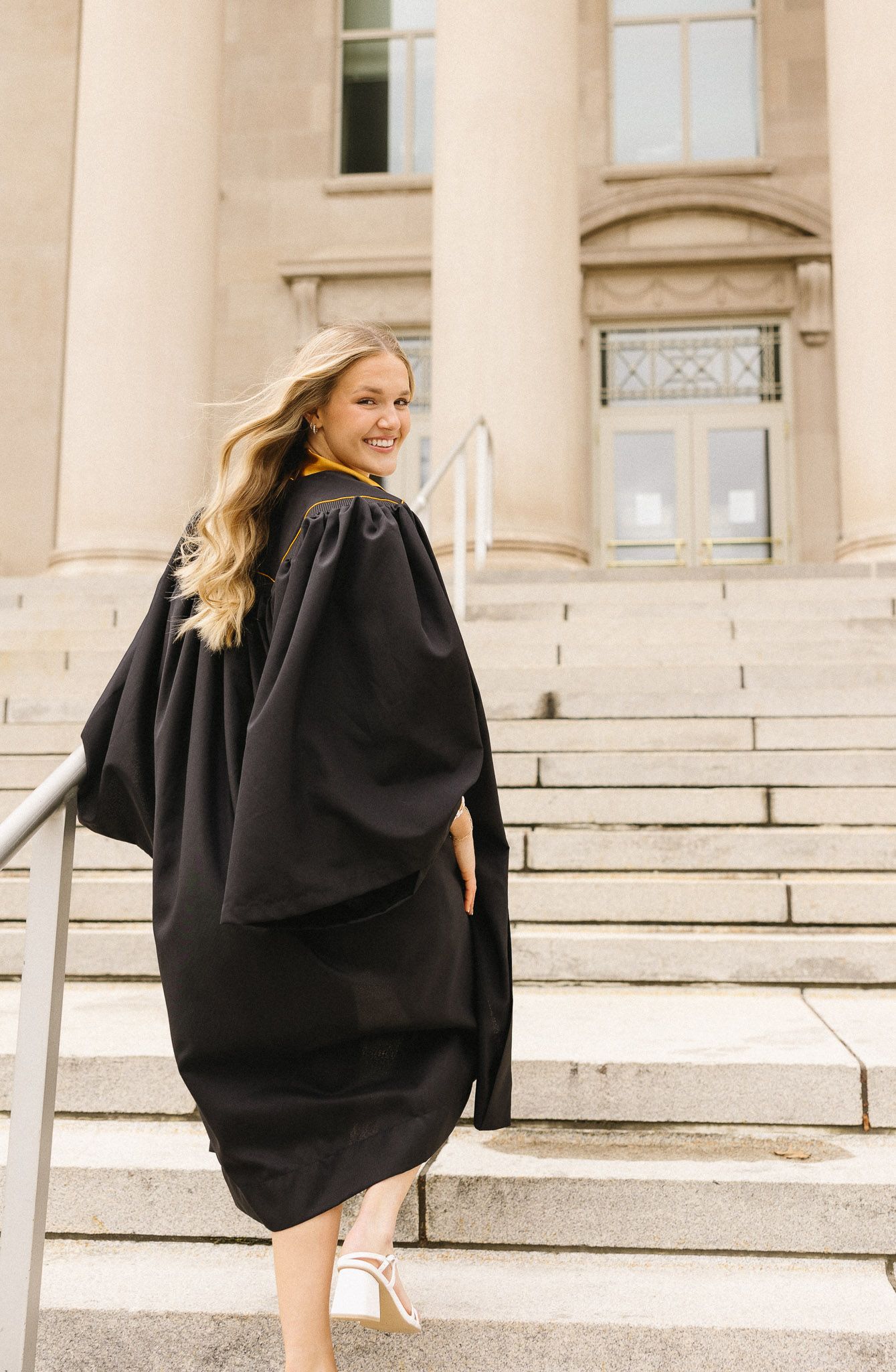 Graduate in flowing black graduation gown poses on stone steps of academic building.