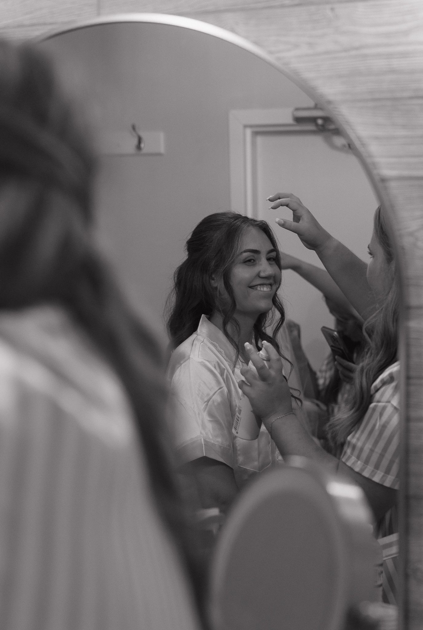 Black and white image of a woman getting ready while looking in a bathroom mirror.