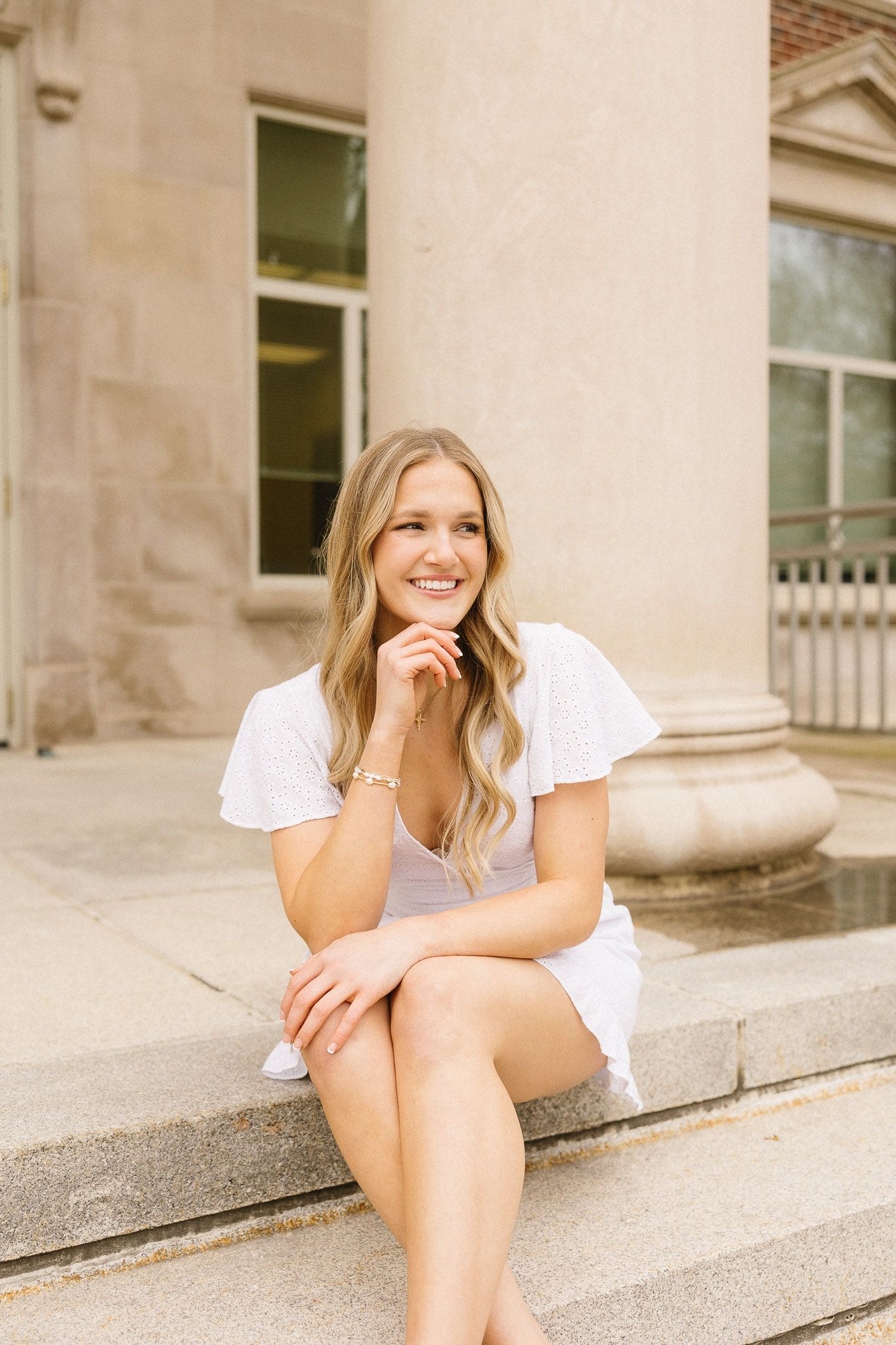 Graduate in white dress sits on stone steps of classical building column.