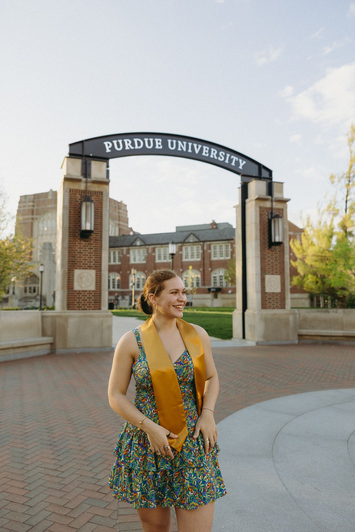 A graduate in a floral dress and gold stole stands in front of the iconic Purdue University arch on campus.