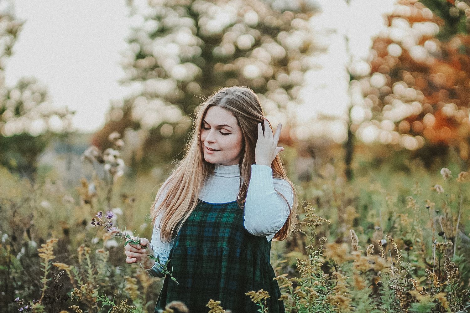 Person in green overalls and white sweater poses thoughtfully in a autumn flower field.