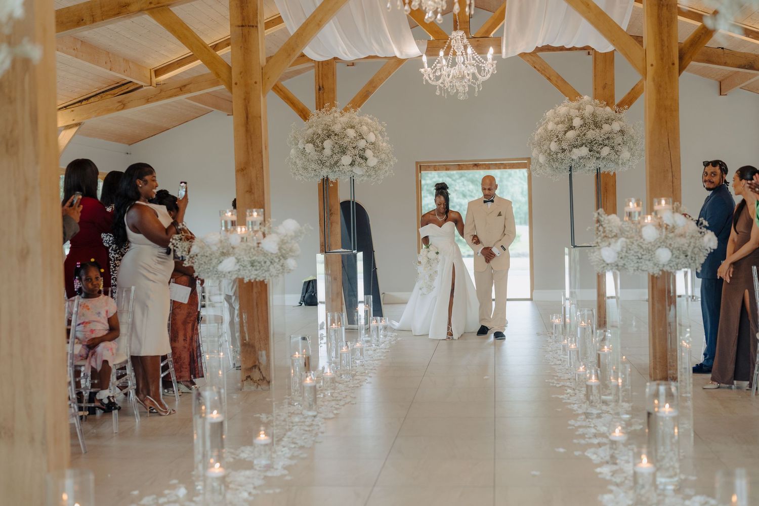 Wedding ceremony in progress in an elegant white room with crystal chandeliers and floral aisle decorations.