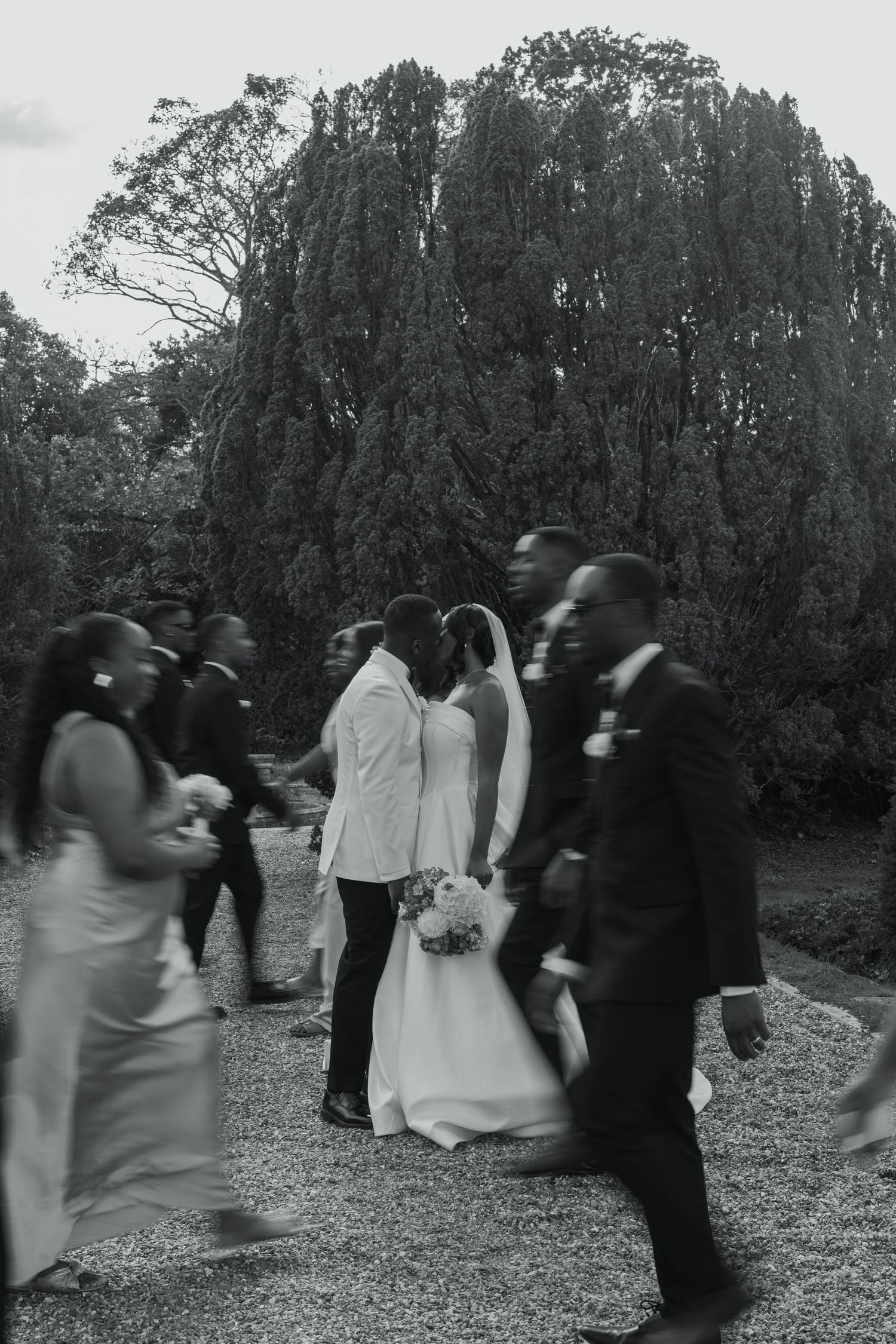Black and white wedding photo of bride and groom kissing surrounded by blurred wedding party guests outdoors.