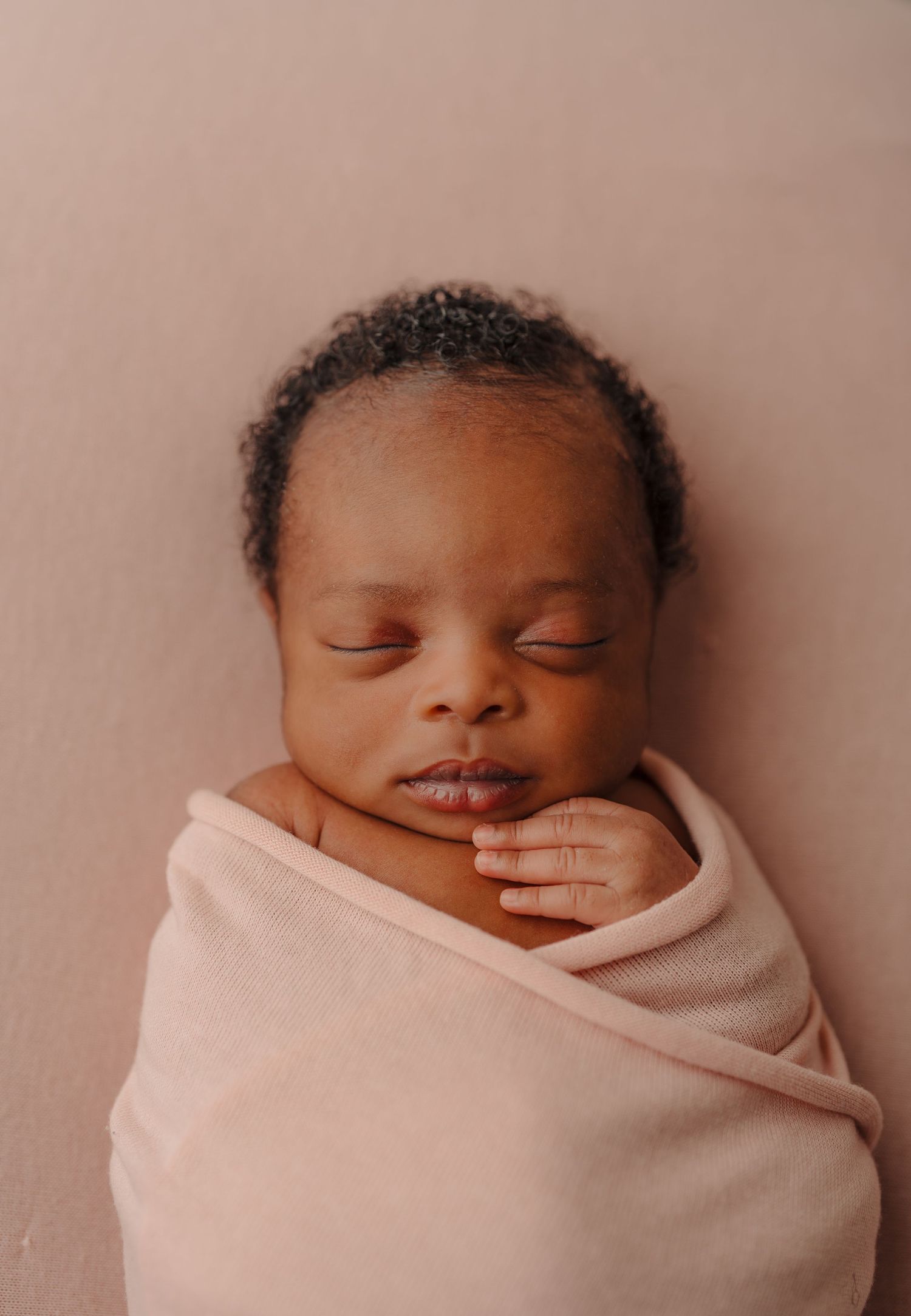 A sleeping newborn wrapped in soft pink fabric against a neutral background.