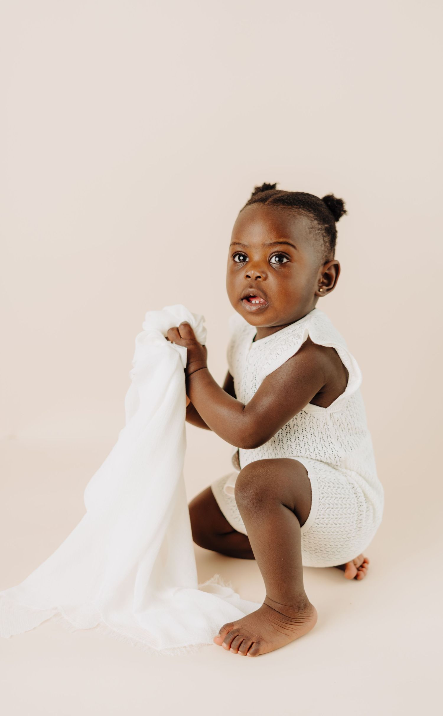 Baby in white knit outfit plays with fabric against neutral background.