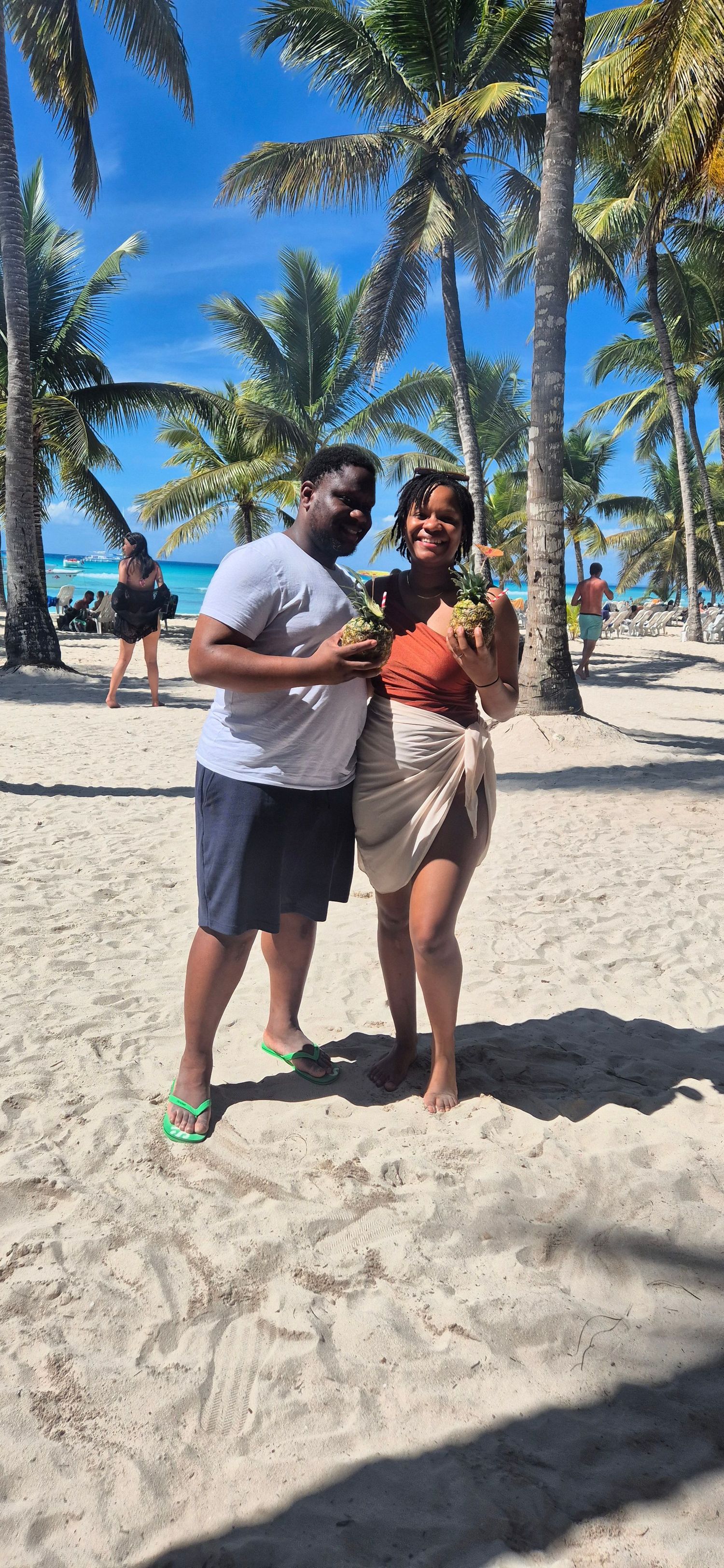 Two people pose playfully on a tropical beach with palm trees and white sand in the background.