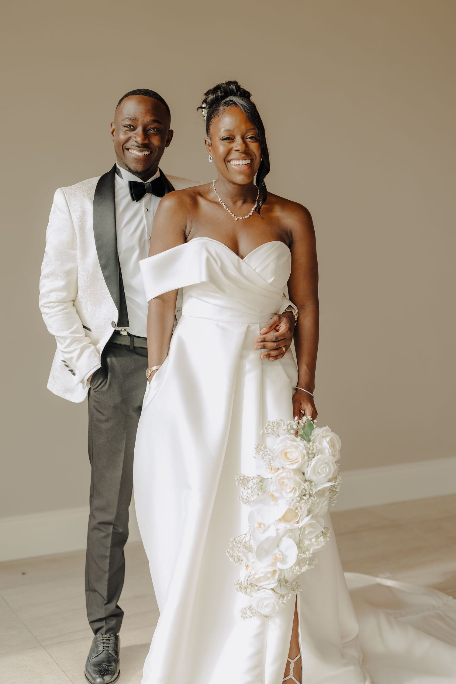 A joyful wedding couple in formal attire poses against a neutral studio background.