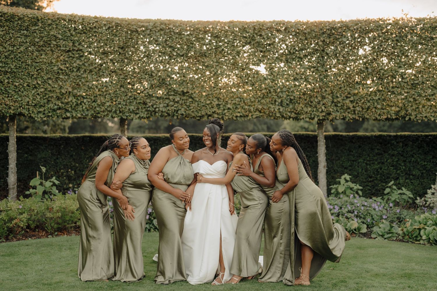 Wedding party members pose together on manicured lawn with greenery in background.