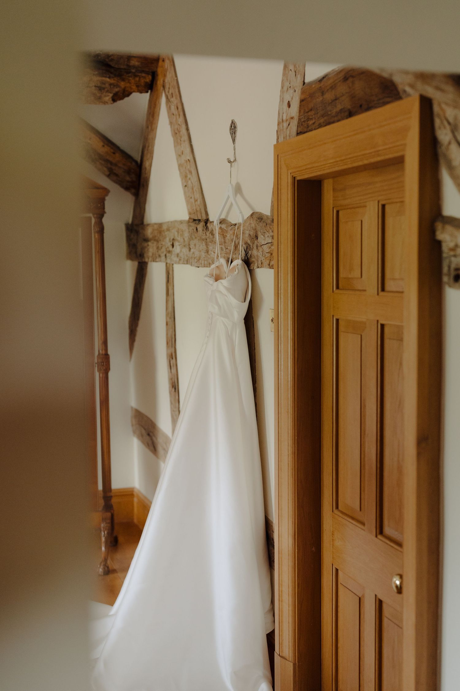 A white wedding dress hangs elegantly against a wooden door and wall in natural lighting.