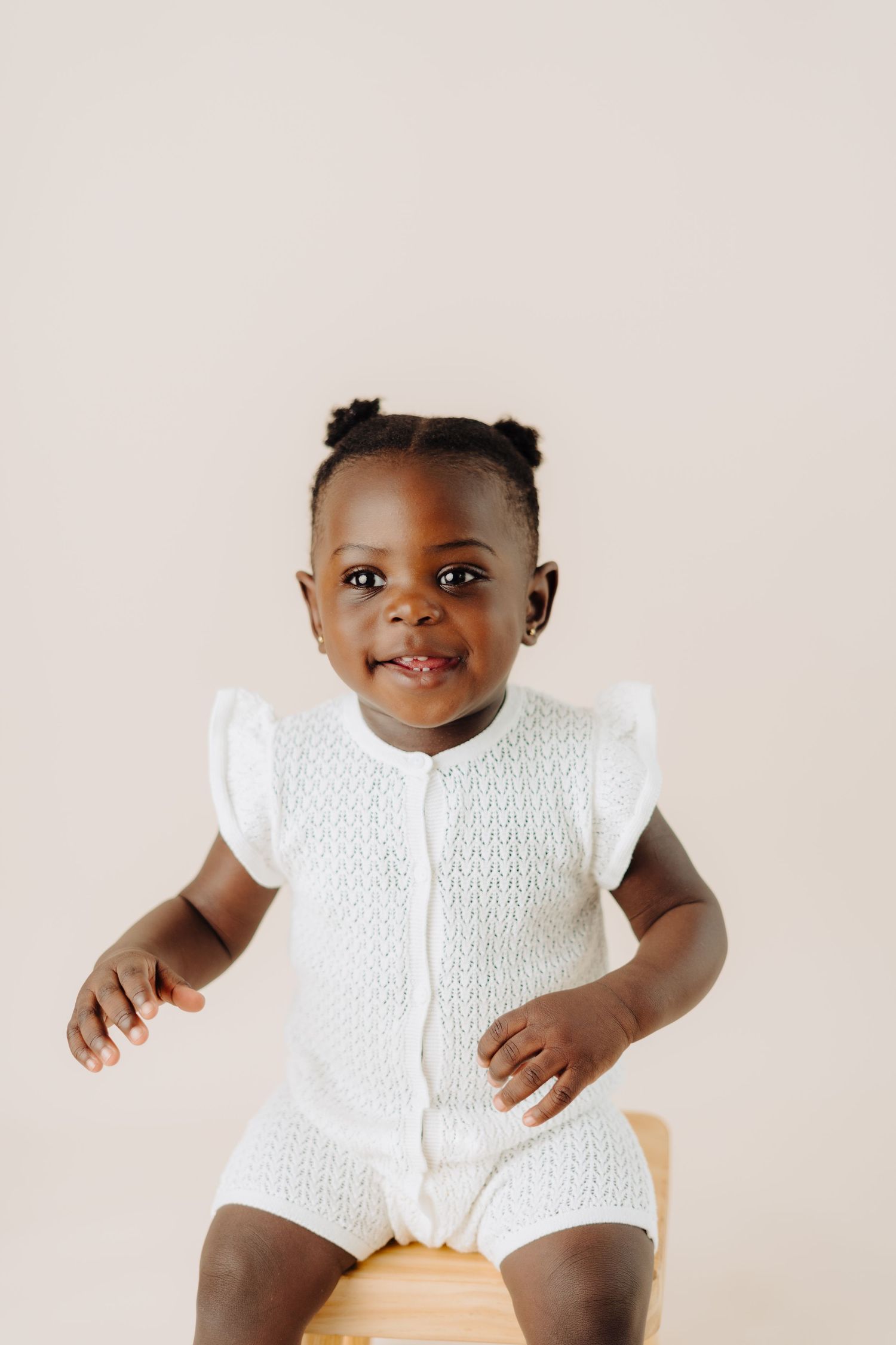 Baby wearing white knit romper smiles joyfully against neutral background.