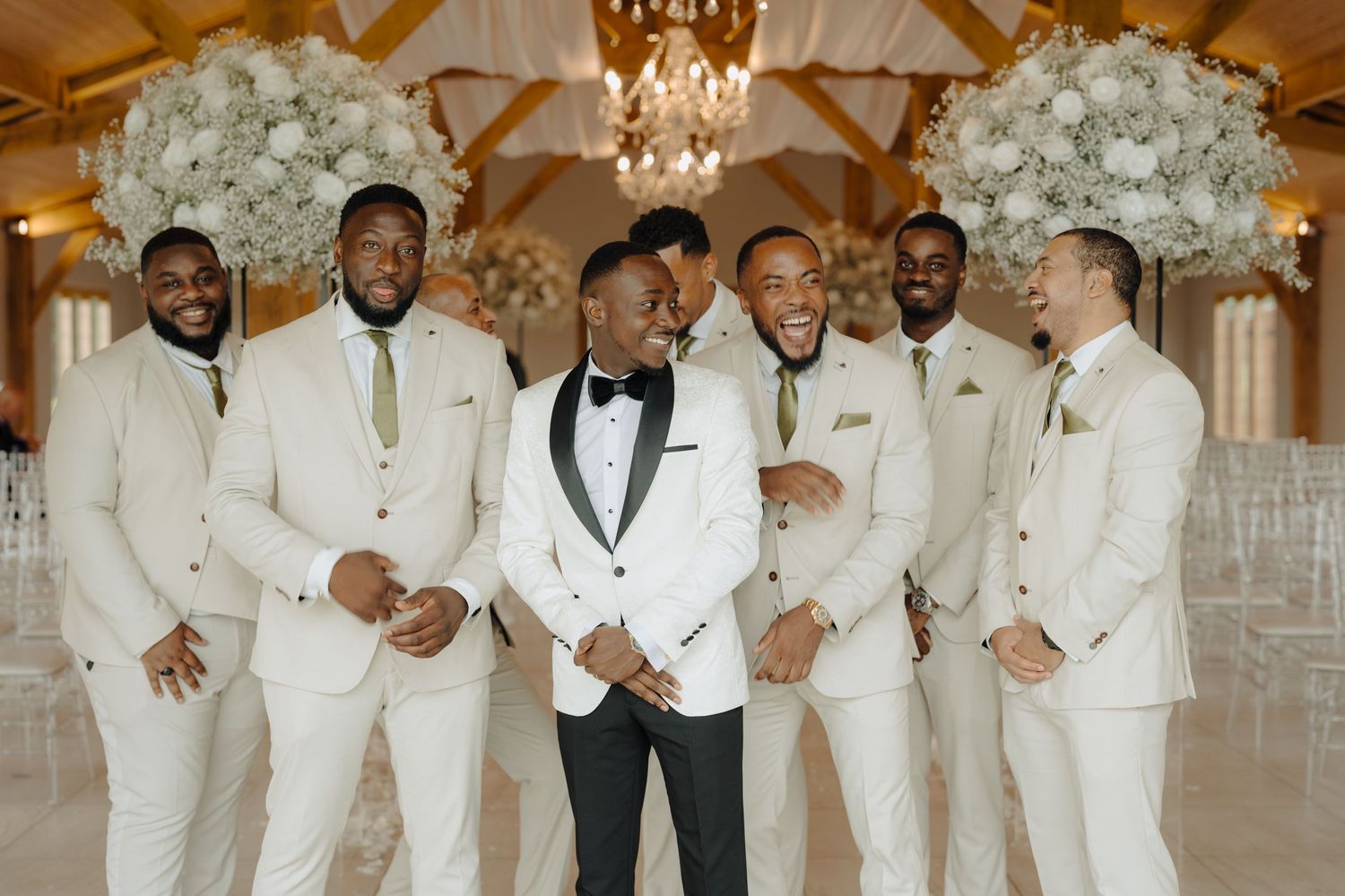 Wedding party members in white suits gather under a chandelier in an elegant indoor venue.