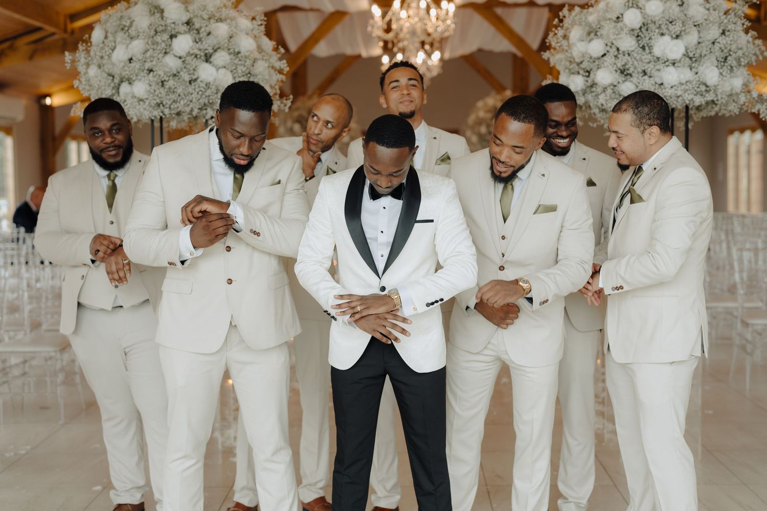 Groomsmen in crisp white suits pose together at an elegant wedding venue with chandelier and floral decor.