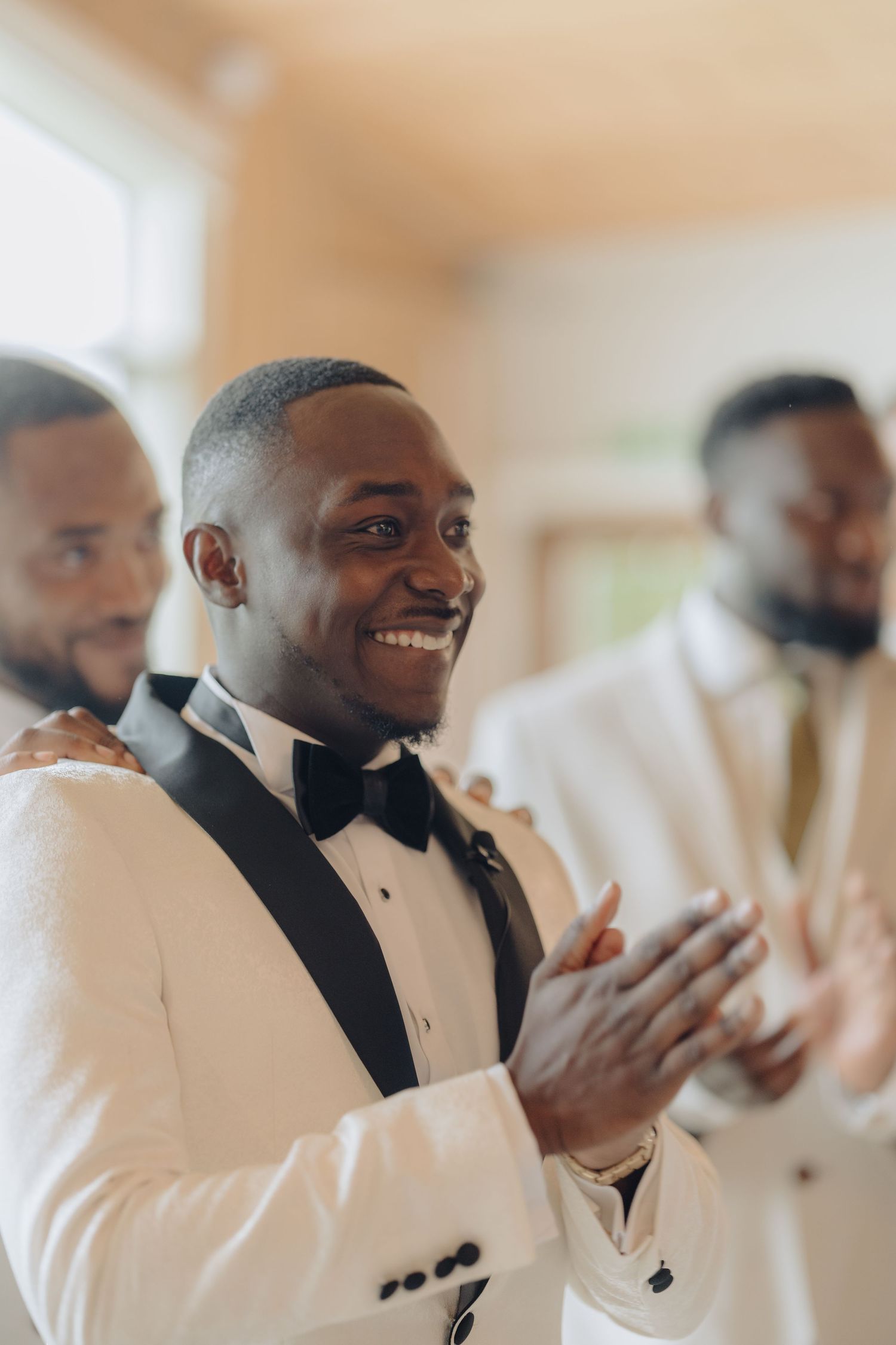 Groomsmen in white tuxedos and black bowties share an emotional moment during wedding preparations.
