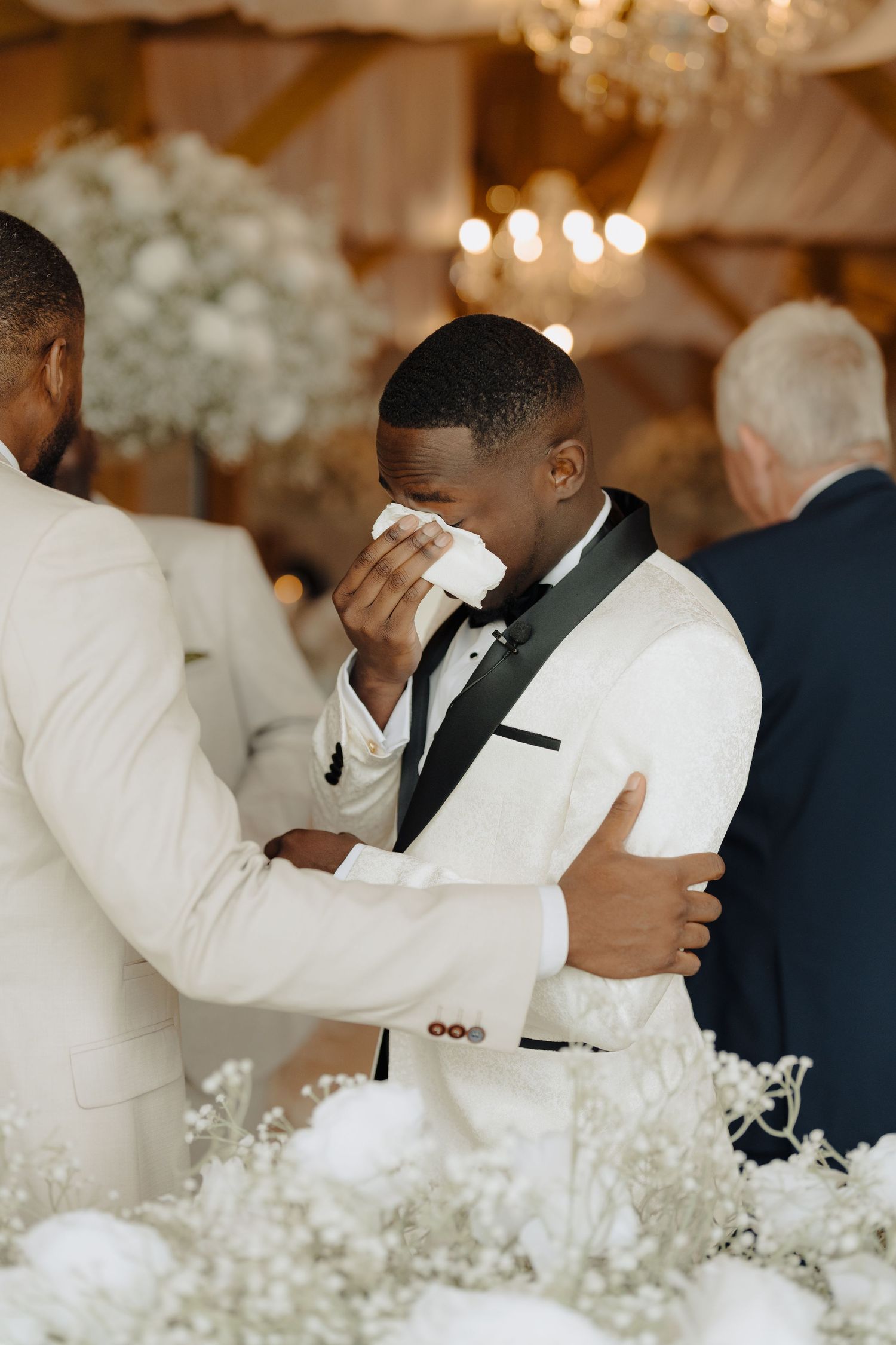 Emotional moment captured during wedding ceremony with white floral decorations and chandelier lighting.