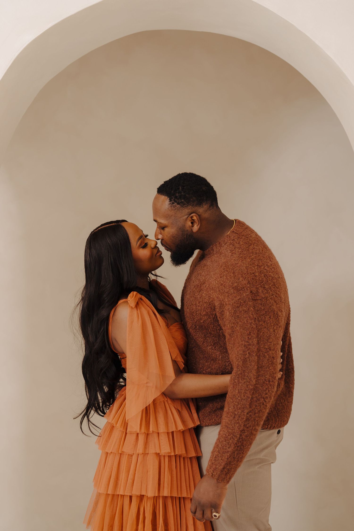 A romantic couple shares intimate moments in an orange dress and brown sweater against a neutral background.