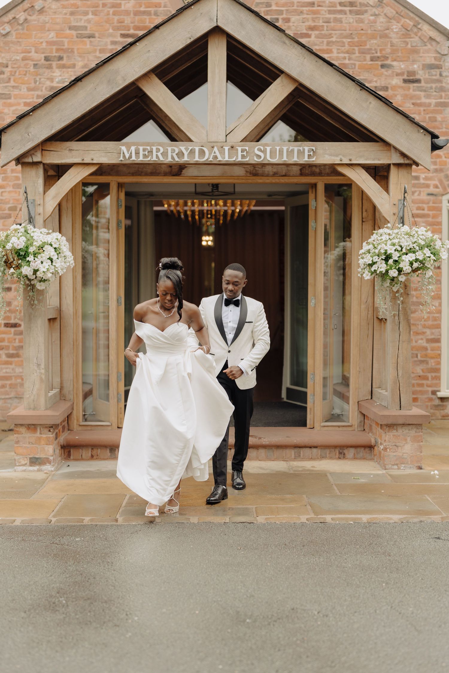 Elegant couple in formal wedding attire exiting a brick building with white floral decorations at Merrydale Suite.