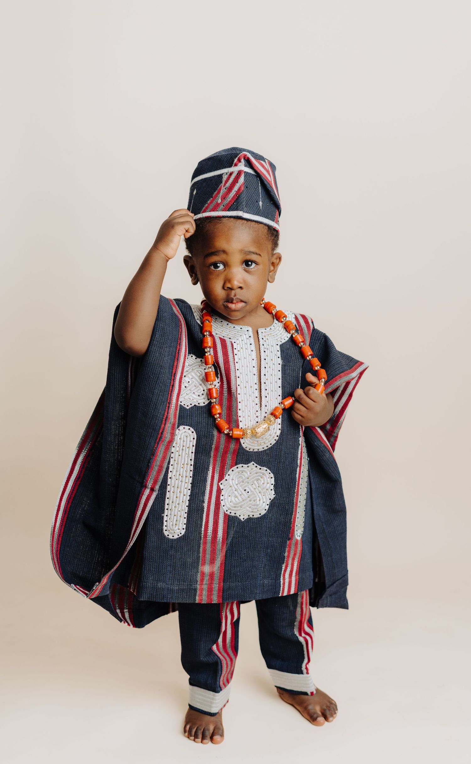 Young child in traditional African clothing and cap stands against neutral backdrop.