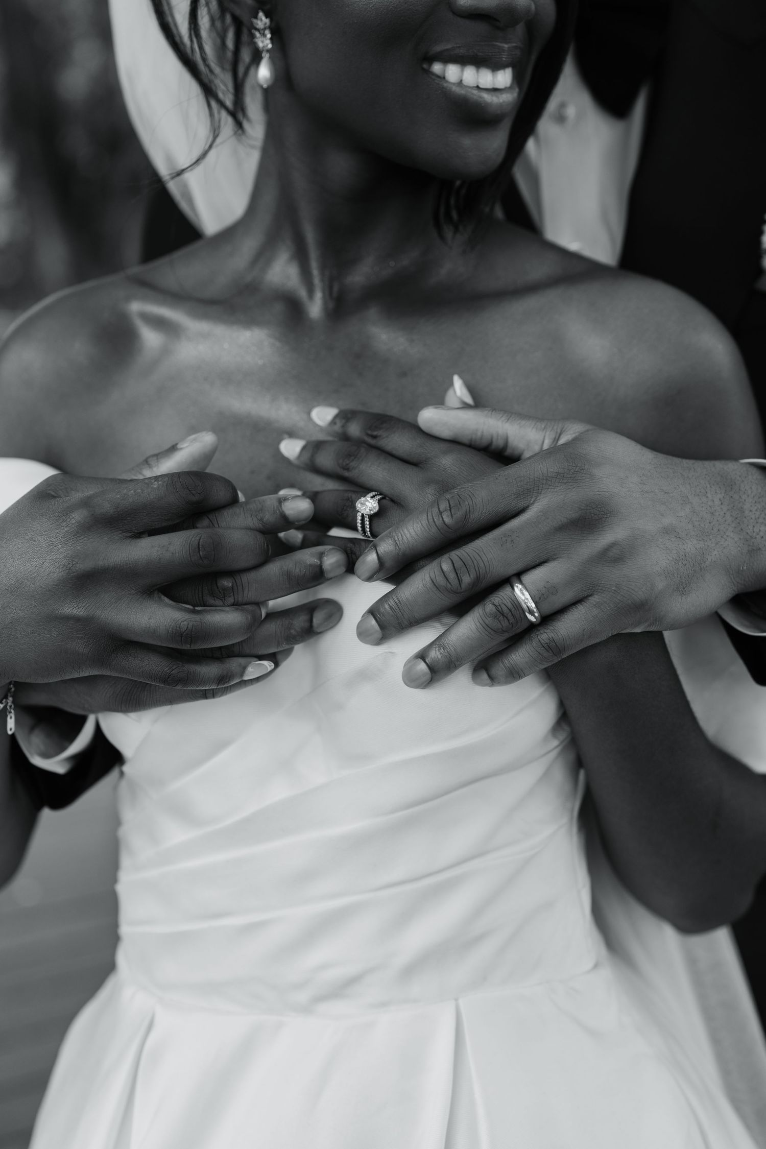 Black and white photo of newlyweds showing wedding rings, bride in strapless gown smiling as groom embraces her.