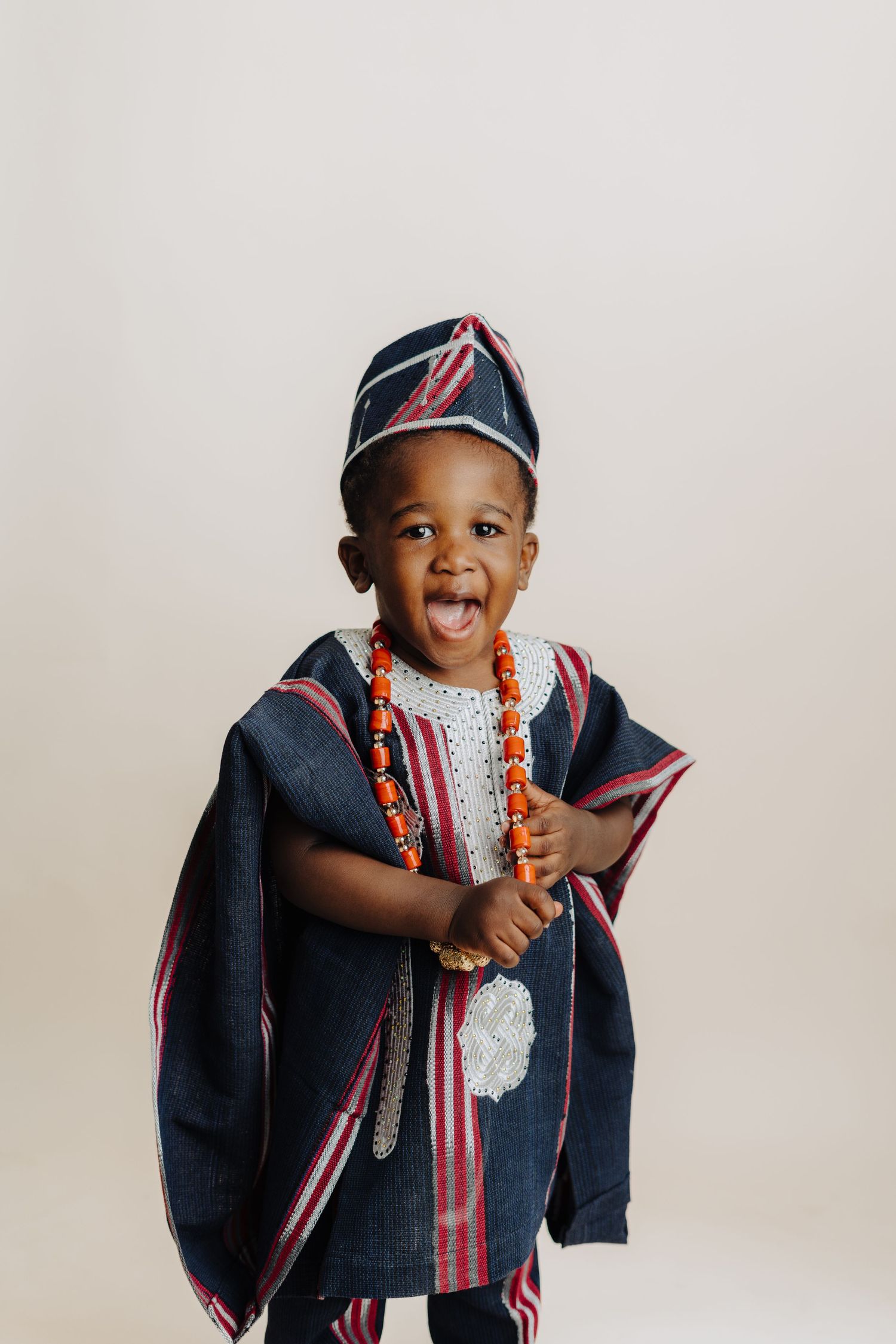Child wearing traditional African attire with cap and beaded necklace poses expressively.