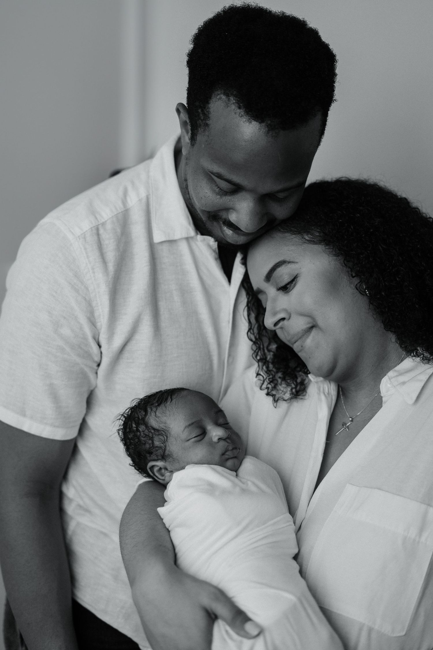 A couple in white clothing shares tender moments with their newborn in a black and white portrait series.
