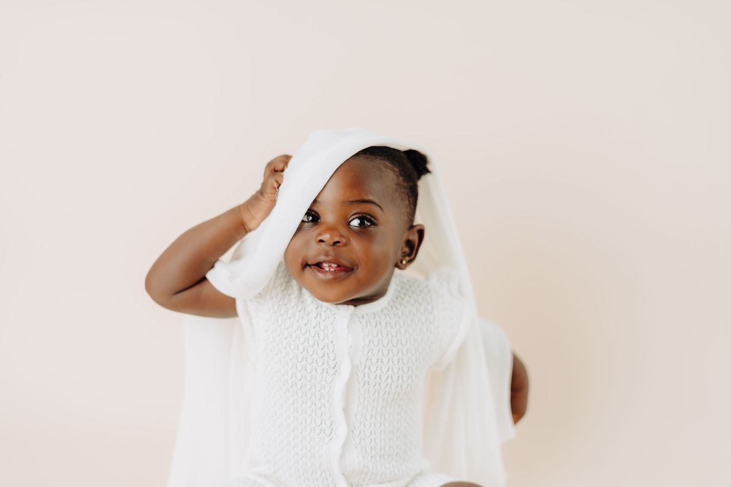 Toddler in white knit outfit raises hand playfully while smiling.