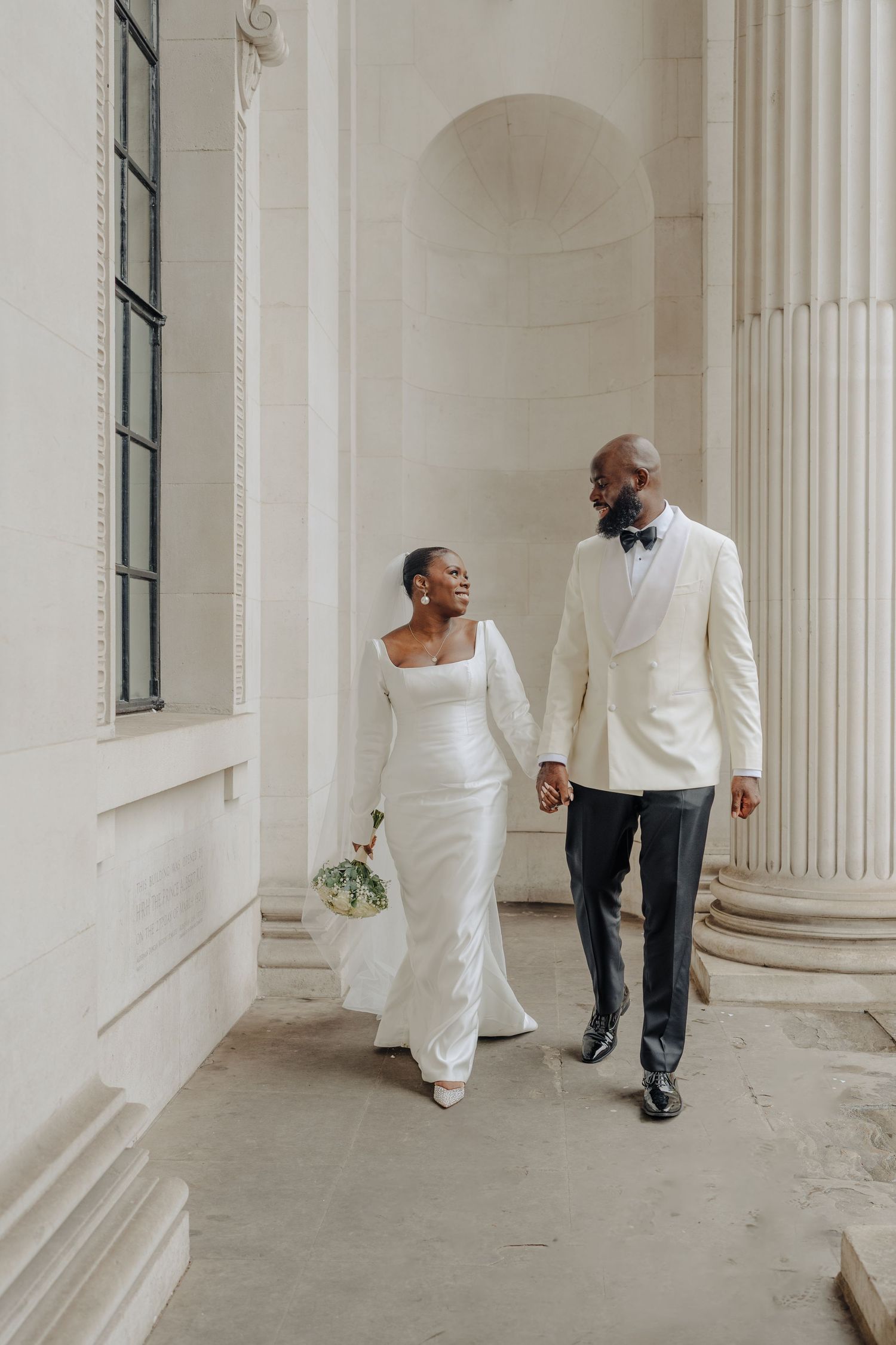 A couple in elegant white attire walks through a columned hallway.