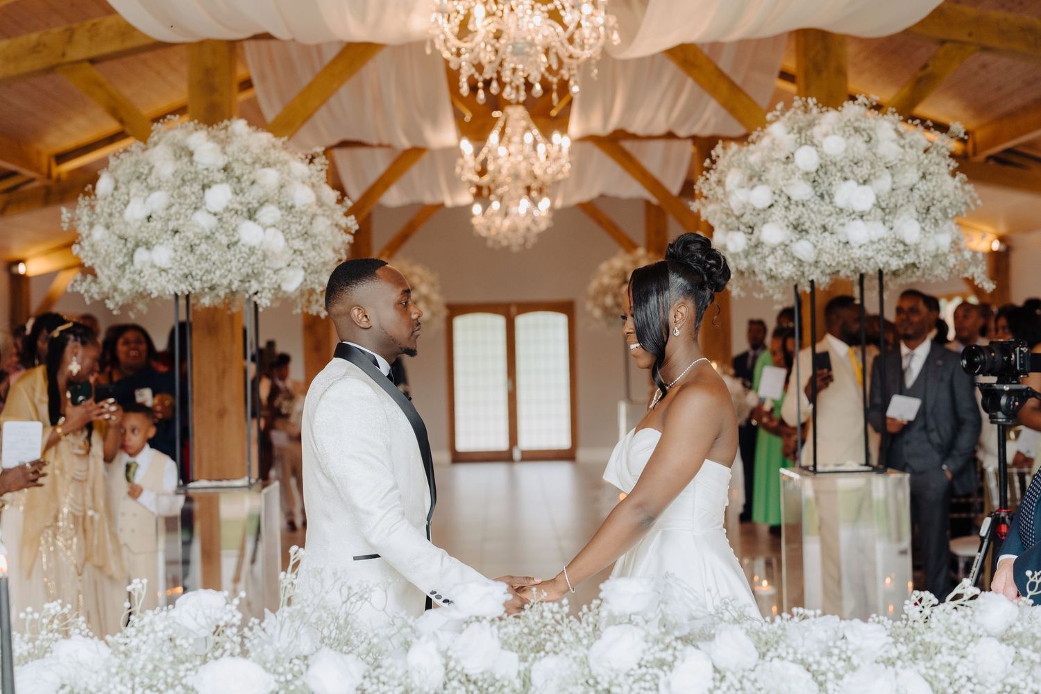 Wedding ceremony with white floral arrangements and crystal chandeliers in a rustic-elegant venue.