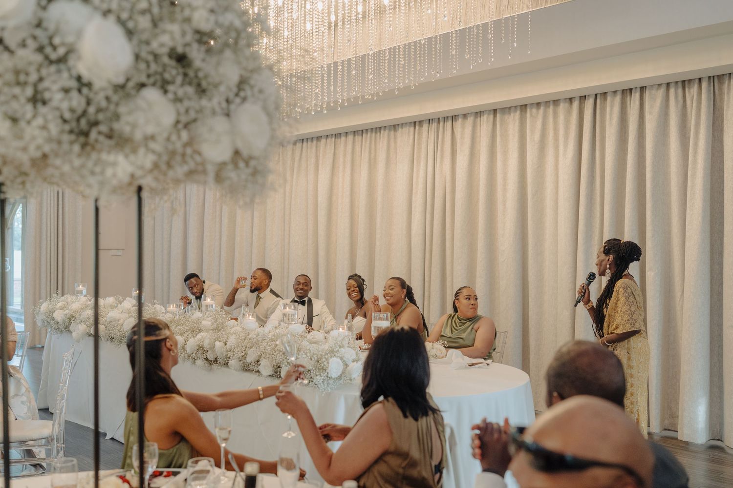 Wedding reception guests seated at elegantly decorated tables with floral centerpieces and crystal chandeliers.