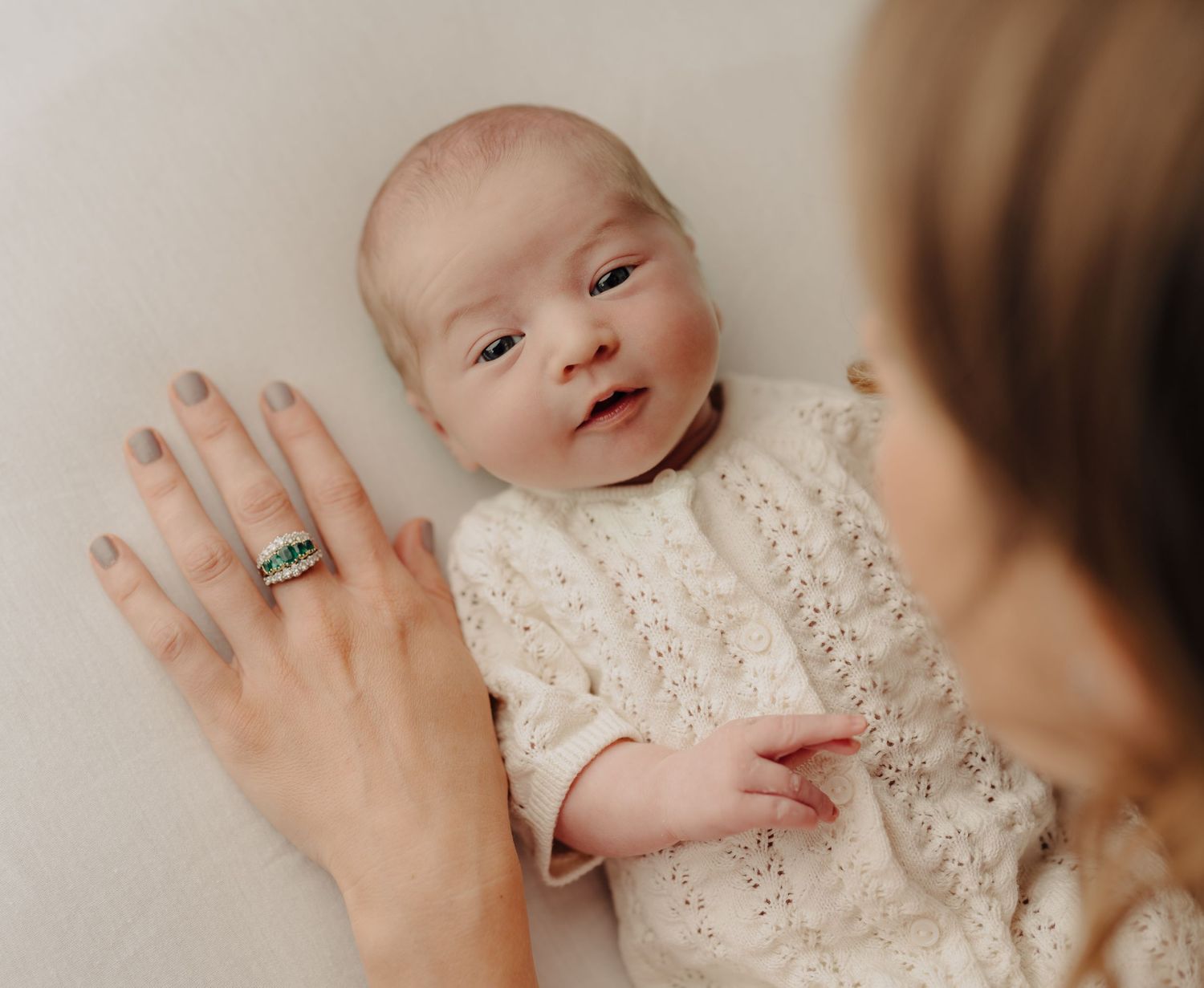 A baby wearing a white lace dress is held by someone wearing a ring.