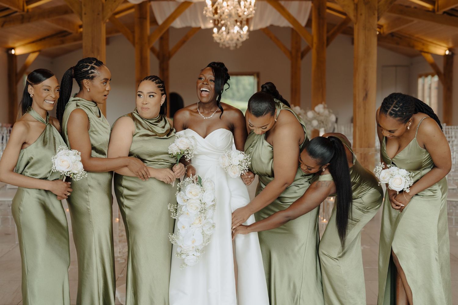 Wedding party in sage green dresses and white gown pose together under wooden beams and chandeliers.