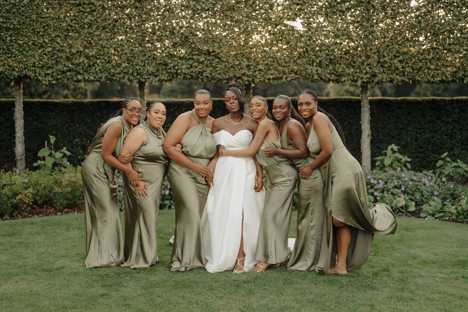 Wedding party in sage green dresses and white gown gather for outdoor photos on landscaped grounds.