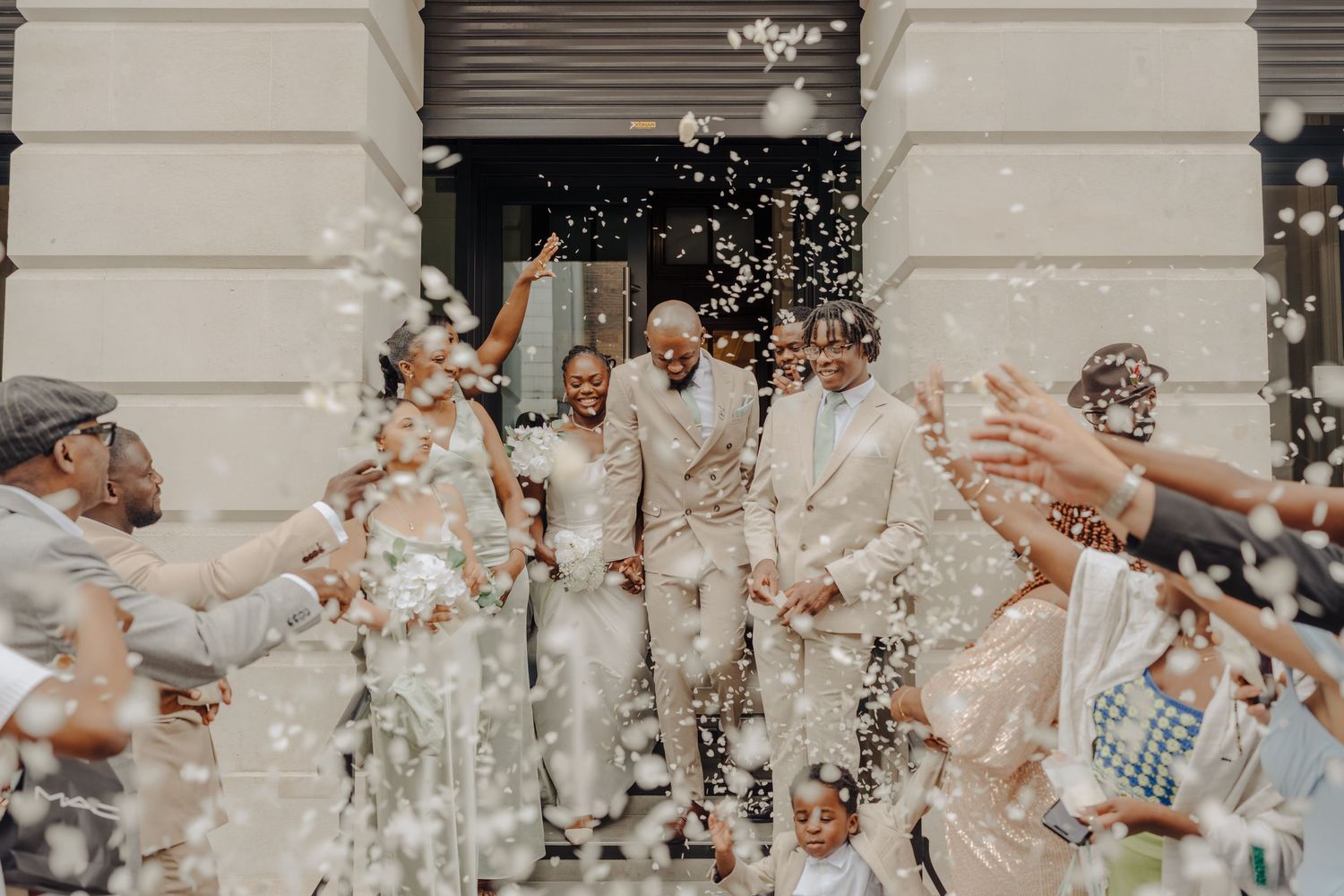 Joyful wedding couple exits venue surrounded by guests throwing white confetti in a bright outdoor celebration.