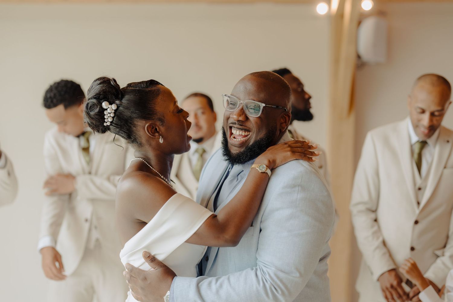Wedding party members embrace and laugh together in a bright indoor venue.