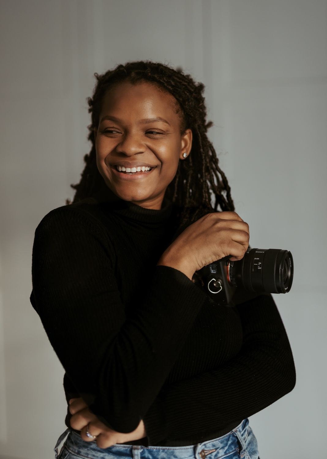 Photographer in black clothing and jeans working with camera equipment against grey background.