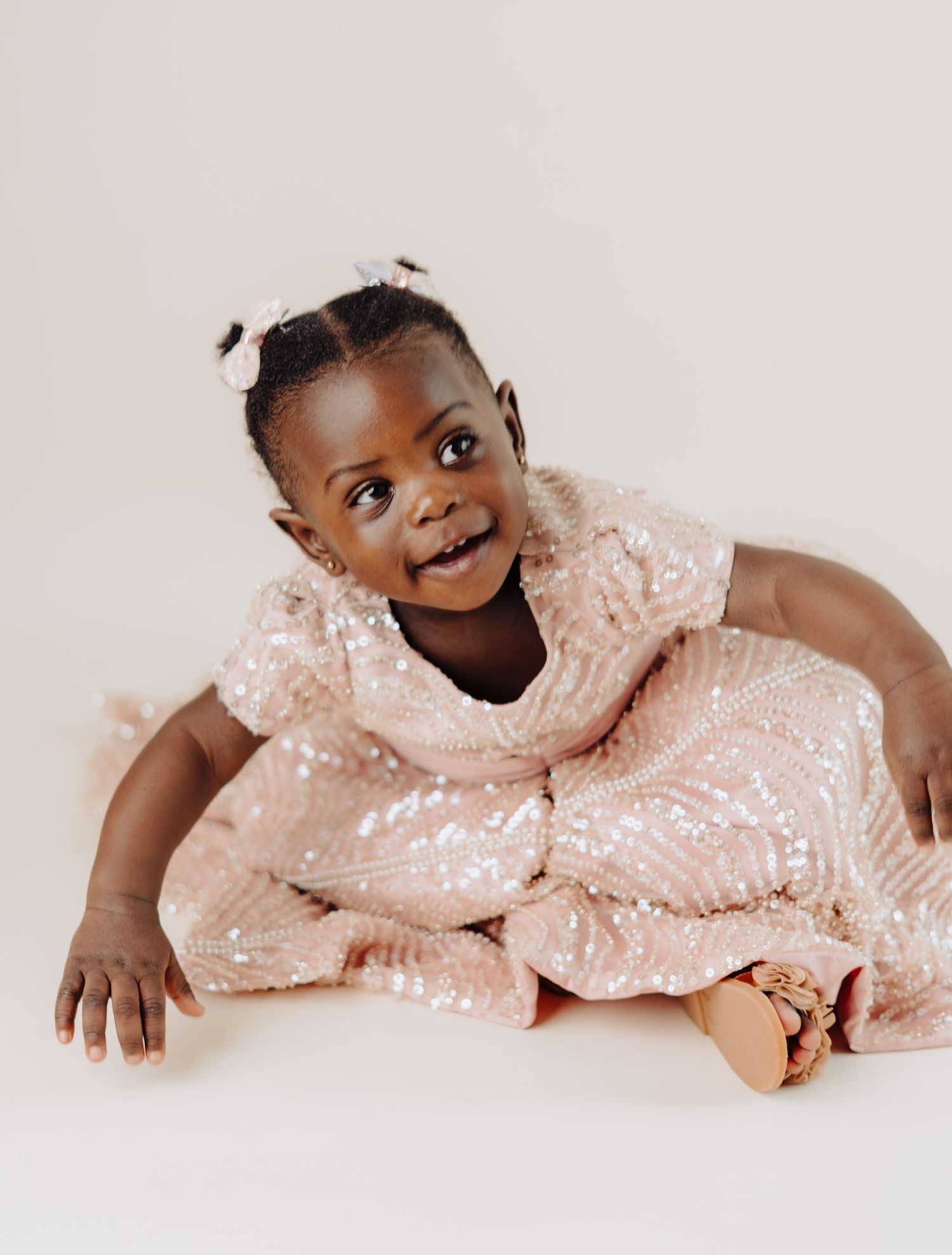 Smiling baby wearing sparkly pink dress sits against light background.