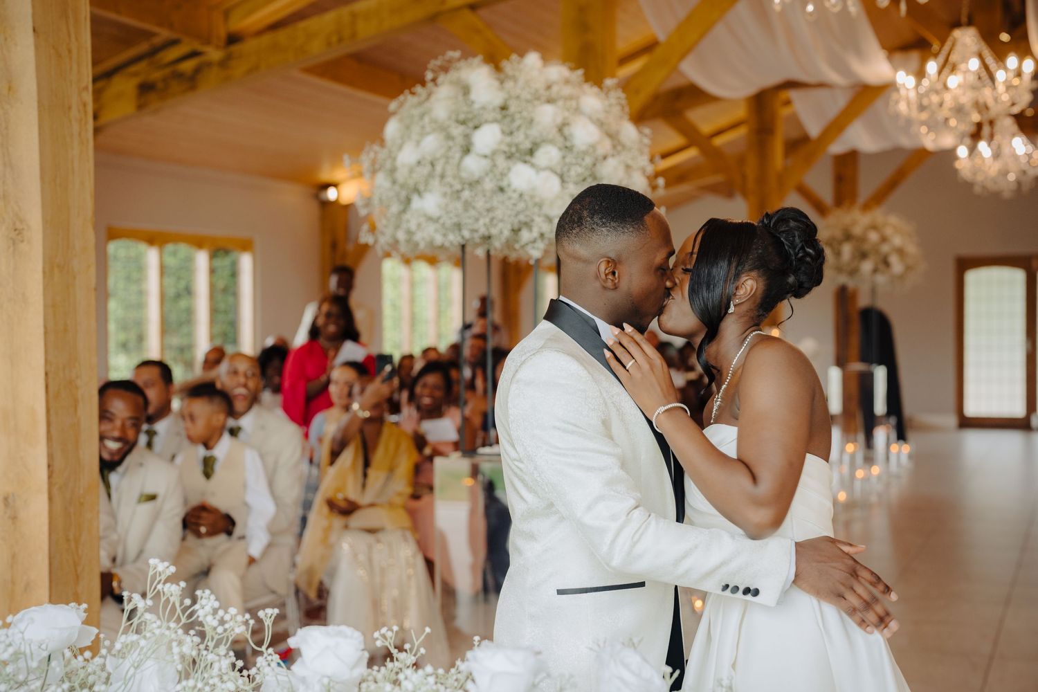 Wedding reception space features exposed wooden beams, crystal chandeliers and white floral decorations.