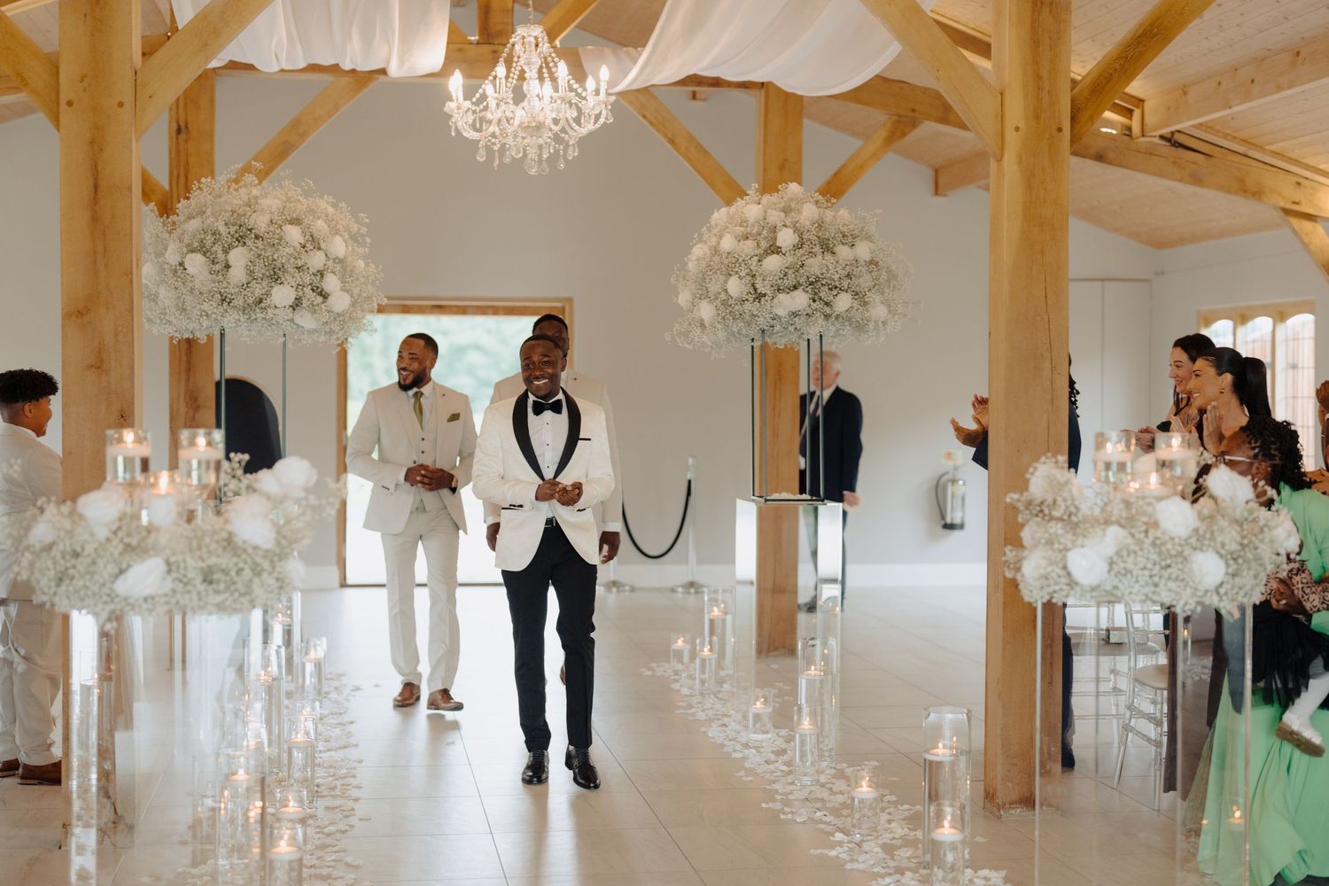 Modern wedding venue interior with exposed wooden beams, chandeliers, and white floral decorations.