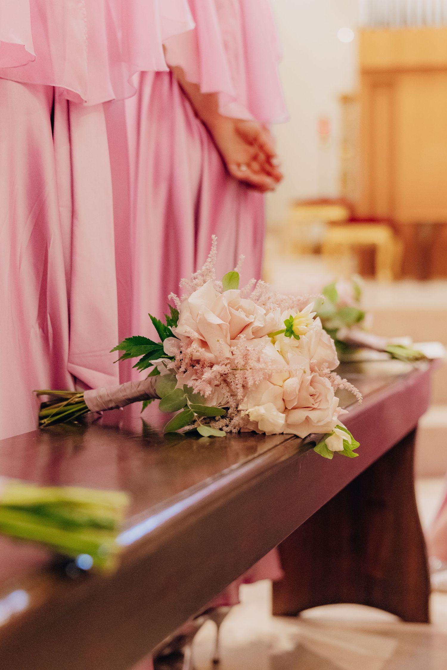 A delicate pink peony bouquet rests on a dark wooden bench beside flowing pink bridesmaid dress fabric.