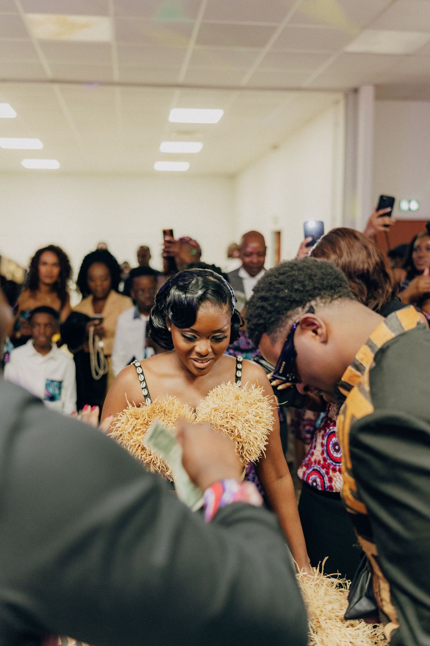 Woman in sparkly gold dress surrounded by celebratory crowd at indoor event.