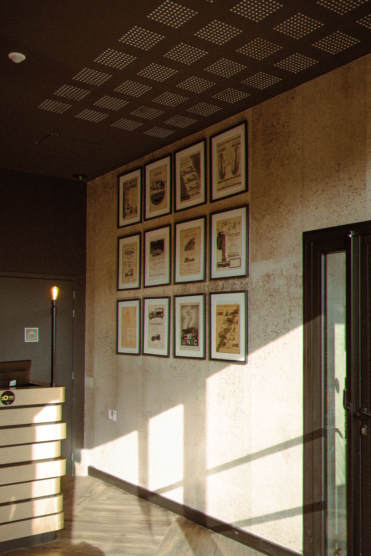 A modern hallway with framed artwork arranged in a grid on the wall, illuminated by dramatic angular sunlight through windows.
