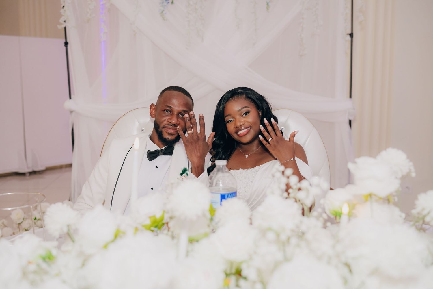 A happy newlywed couple shows off their rings while posing behind white floral decorations at their wedding reception.
