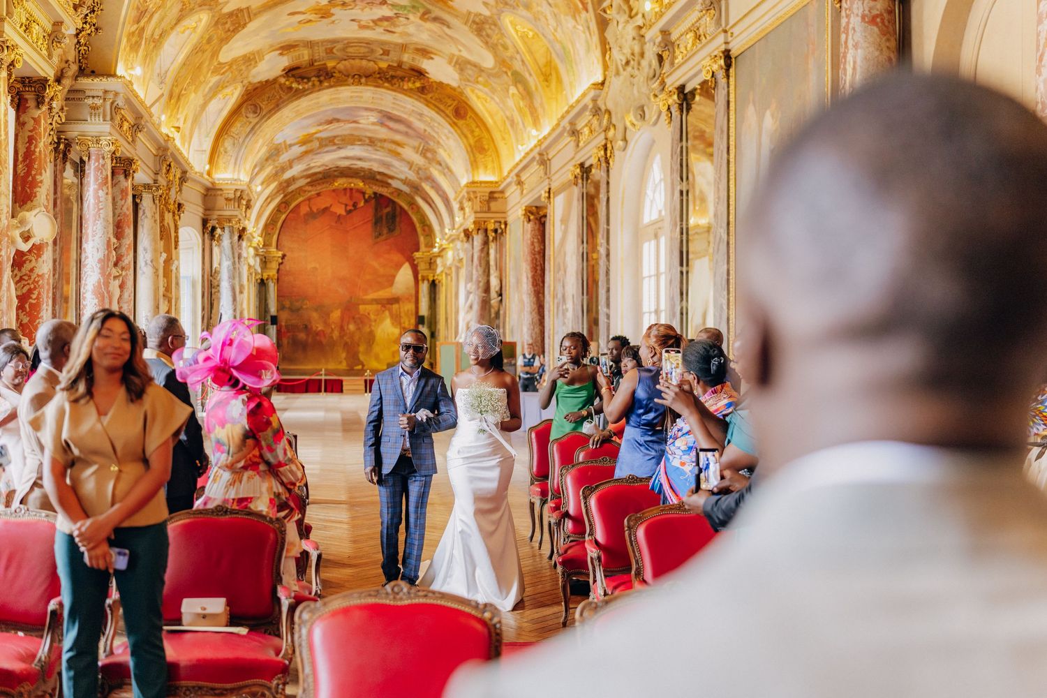Wedding ceremony in an ornate palace ballroom with gilded baroque architecture and red carpeting.
