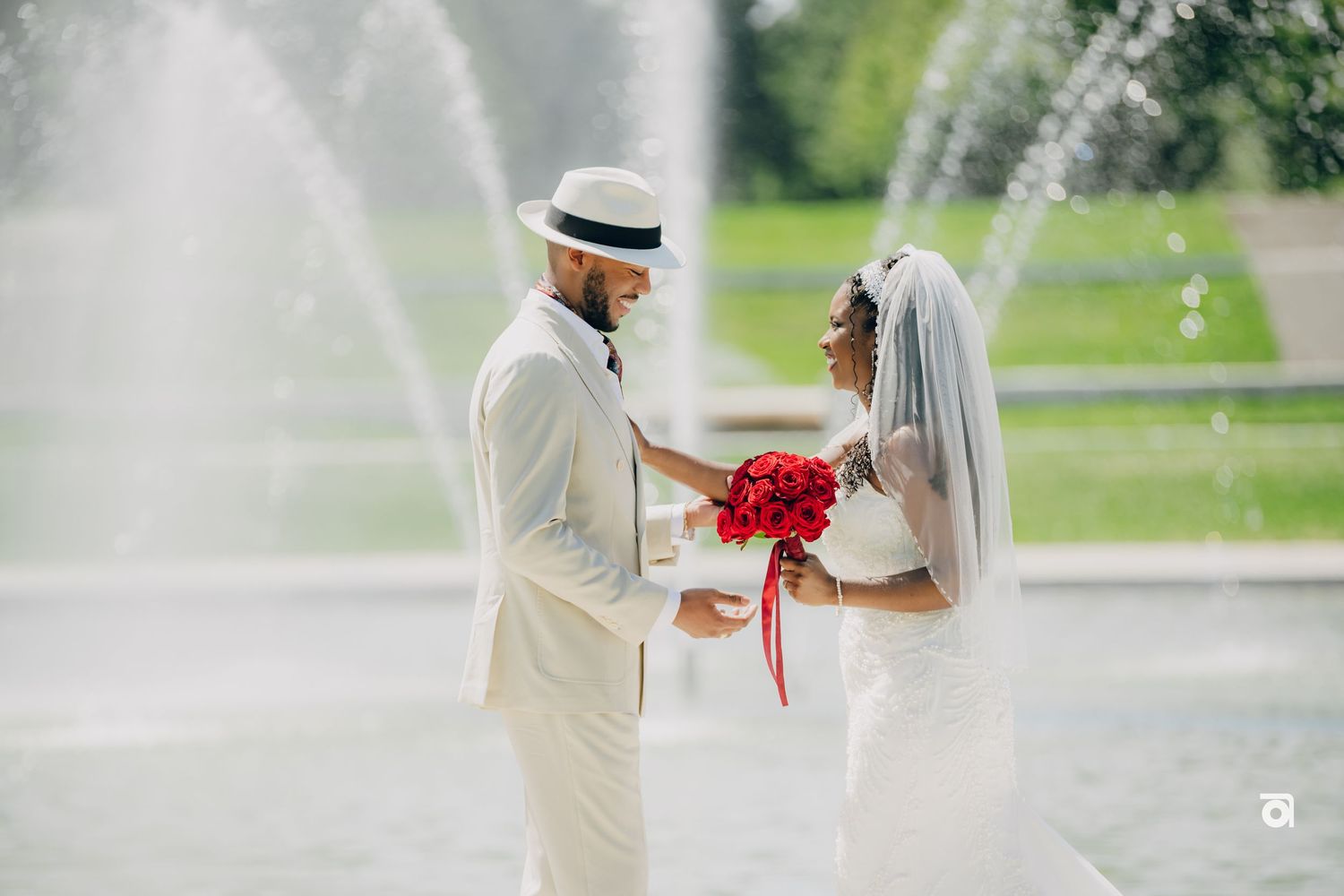 A couple in white wedding attire holds hands in front of a fountain spray in a garden setting as they exchange vows.