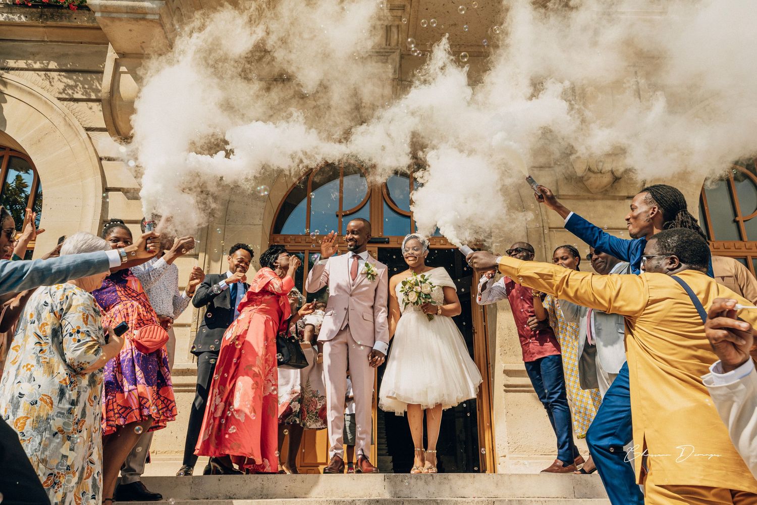 Wedding celebration with smoke effects and guests in colorful attire outside historic building.