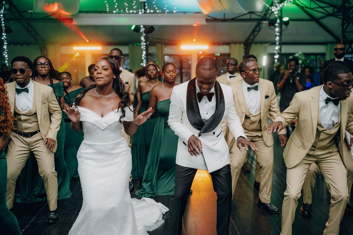 Wedding party dancing in coordinated green and beige outfits under colorful lighting.