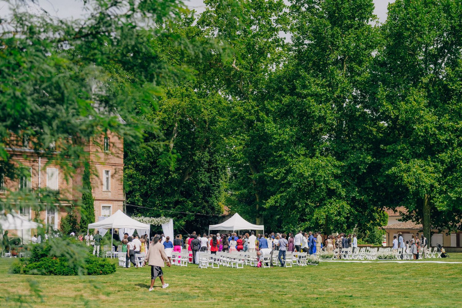 Wedding guests gather on a green lawn near historic brick buildings with white tents and umbrellas set up outdoors.