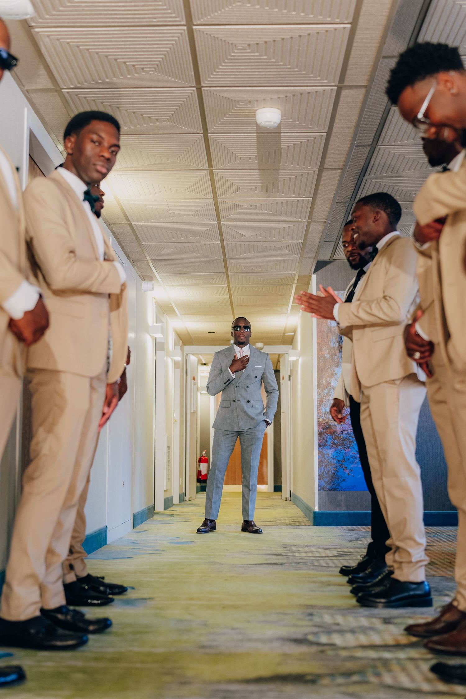 A group in matching beige suits poses in a hallway with one person in gray standing in the center.