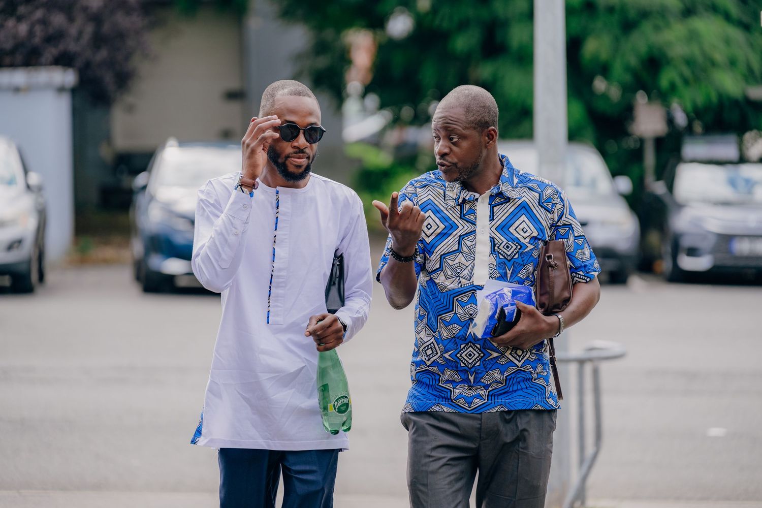 Two people walking and conversing together outdoors, one wearing a white shirt and another in blue pattern.