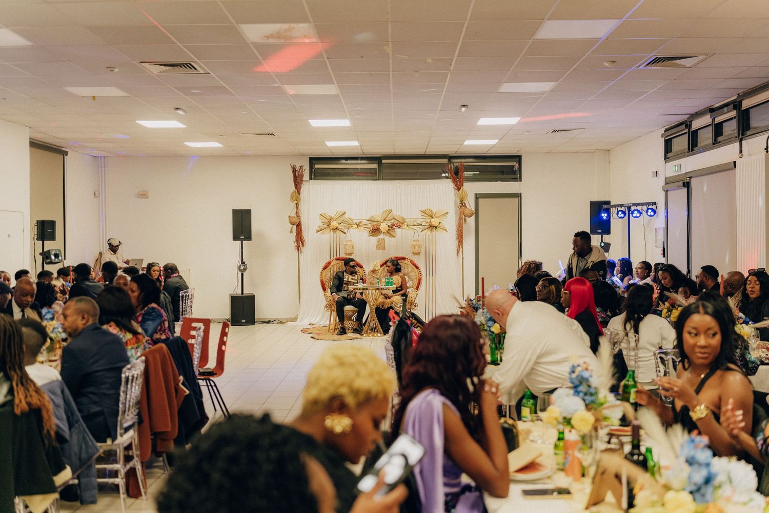 Wedding reception guests seated at decorated tables in a banquet hall with a stage and floral arch setup.