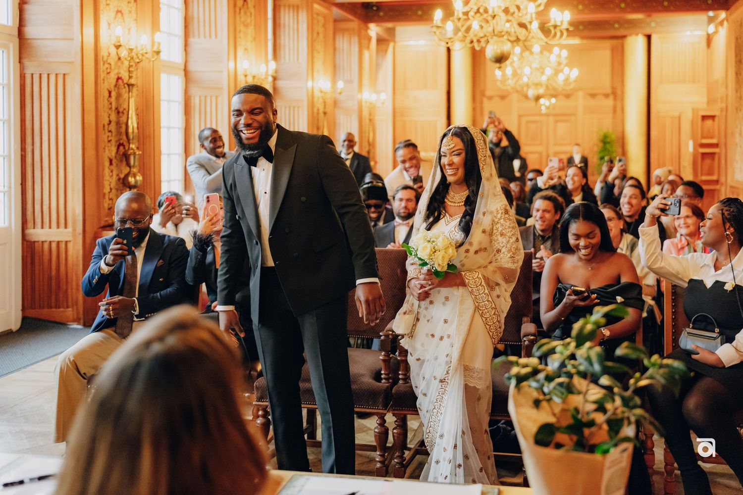 Newlyweds share joyful moment during wedding ceremony in elegant ballroom with chandelier and floral arrangements.